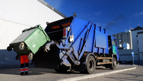 A large blue refuse collection vehicle with an open rear compartment is engaged in rubbish collection on a paved urban street. The truck's rear hopper is lifted, revealing the interior where waste materials are being deposited. To the left, a person wearing bright orange high-visibility trousers and dark boots stands next to a green wheeled waste bin, which is partially lifted and positioned close to the truck's opening. The bin has a textured plastic surface and is tilted slightly forward, with the lid closed. The background features a white industrial or commercial building with vertical paneling and a flat roof, set against a clear blue sky. Nearby provides a sense of an organized waste management operation, consistent with professional rubbish removal services, which might be used by residents or businesses in Leatherhead or the surrounding area. The scene is well-lit by natural daylight, with shadows indicating the sun is relatively high in the sky, emphasizing the activities associated with waste collection in a typical urban environment.