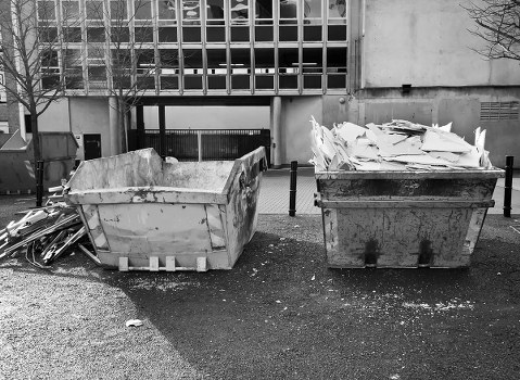 Two large industrial dumpsters are positioned side by side in an outdoor area, with the left container filled with irregularly shaped, broken concrete slabs featuring rough textures and light grey surfaces. The right container is overflowing with flattened cardboard boxes and paper waste, which appear slightly crumpled and torn, with varying shades of white and off-white. The dumpsters are situated on a paved surface adjacent to a modern building with a grid-like facade composed of glass and metal panels, casting slight shadows around the area. Surrounding the containers, scattered debris and small particles of waste are visible on the ground, with a few metal objects leaning against the left dumpster. In the background, a portion of a concrete wall, some leafless trees, and a security barrier can be seen, indicating a utilitarian environment likely associated with waste collection or rubbish disposal, consistent with services provided by House Clearance Leatherhead in the Surrey area near KT22 postcode. The overall scene depicts a typical rubbish removal setup for handling construction debris and general waste collection. The lighting appears natural, suggesting daytime with overcast skies.