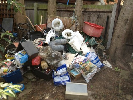 A collection of mixed household waste and rubbish piled on a patch of soil in a garden area, including plastic buckets, cardboard boxes, plastic bags, and various household items, some stacked and leaning against wooden fence panels. The waste appears disorganized, with items such as a gray plastic pipe, a red plastic container, and various smaller objects intermixed. The background shows a weathered wooden fence with vertical slats and surrounding trees or shrubs, indicating an outdoor setting typical of a residential backyard in Leatherhead. The scene is illuminated by natural daylight, highlighting the variety of materials and textures, from smooth plastic to rough cardboard and rough wood. The image aligns with rubbish removal services, such as those offered by House Clearance Leatherhead, supporting the need for clearing accumulated waste from private residential gardens or outdoor spaces.