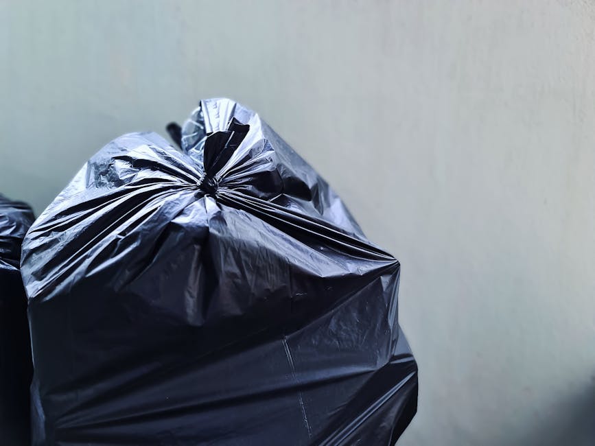 Close-up image of a black plastic rubbish bag secured with a twist tie, placed against a plain, light-colored wall. The bag's glossy surface reflects light, revealing its crinkled texture and the tigh