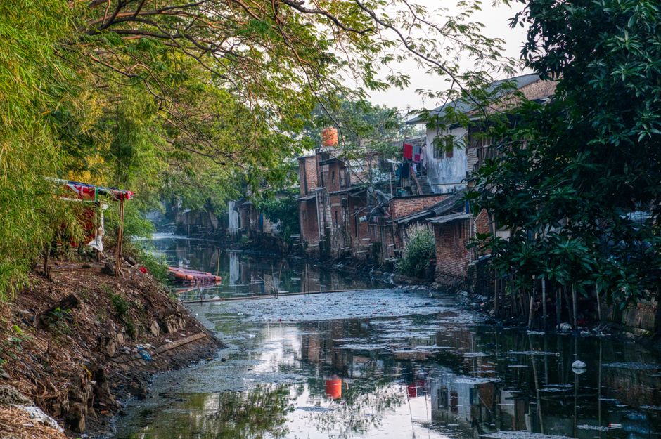 A narrow waterway or canal running through a residential area with buildings constructed from red brick and concrete on the banks, some with visible weathering and minor structural wear. The water's s